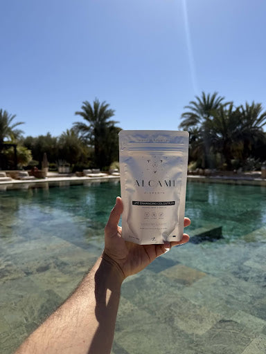 A person holding a packet of Alcami Element’s Life Enhancing Colostrum by a poolside with palm trees in the background.