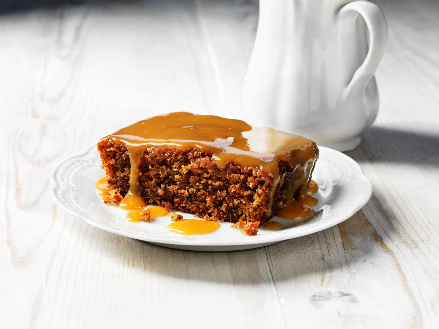 A slice of sticky toffee pudding topped with rich caramel sauce on a white plate.