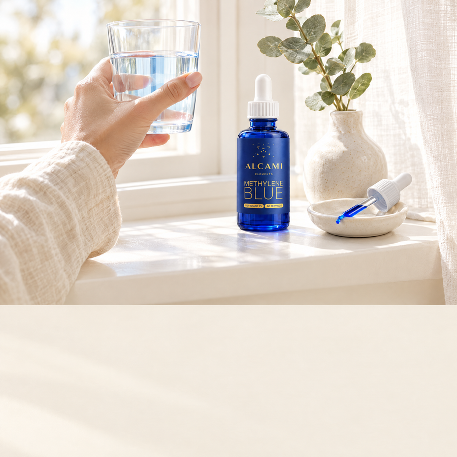 A hand lifting a glass of water by a bright window next to the Alcami Methylene Blue bottle